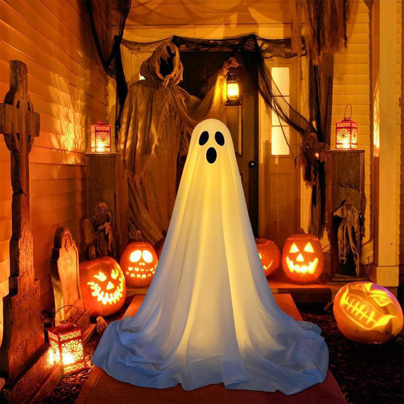 A Halloween decoration featuring a glowing white ghost figure draped over a form, set on a porch surrounded by carved pumpkins, lanterns, and spooky decor.