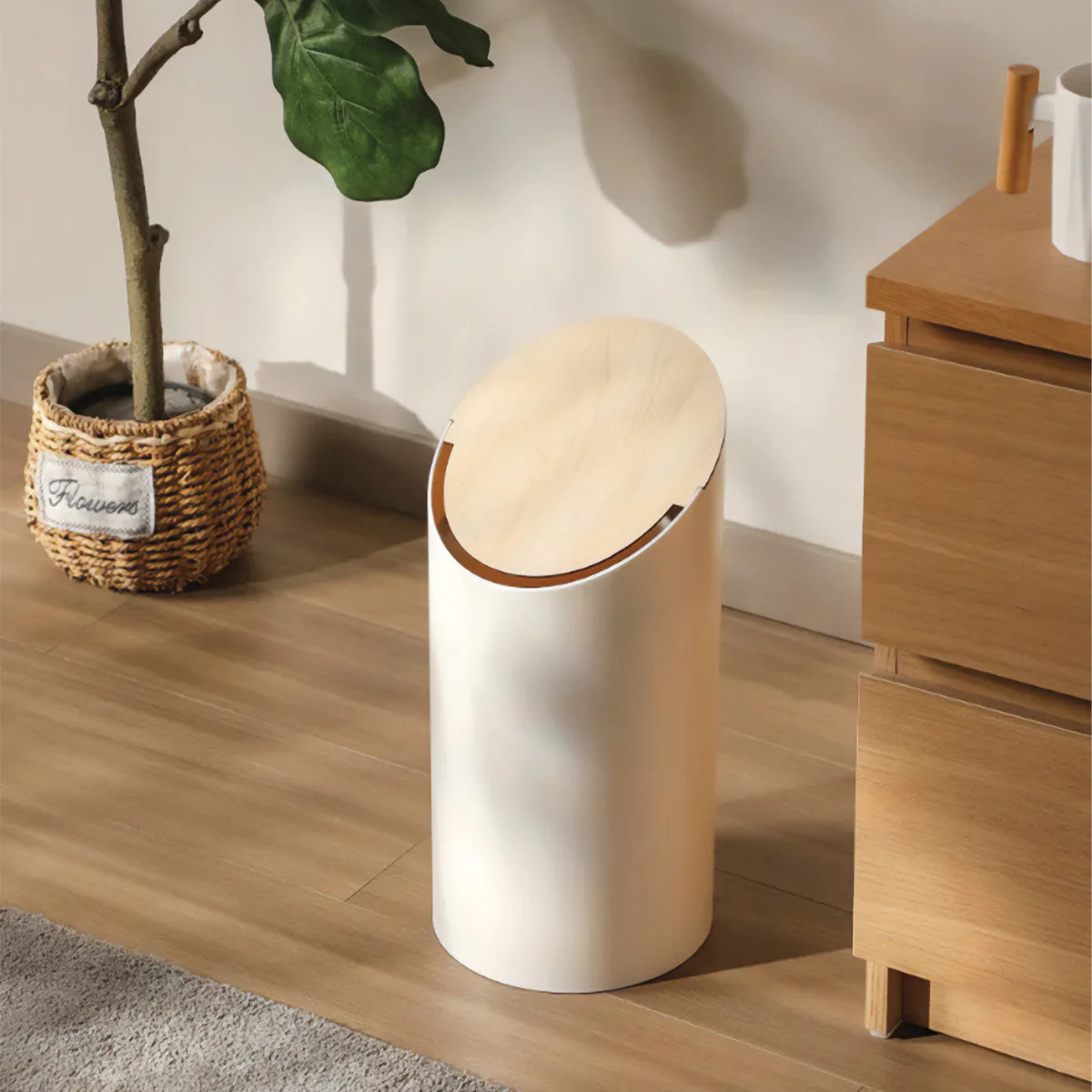 White trash bin with wooden accents on a wooden floor next to a plant and dresser.