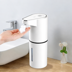 White automatic touchless soap dispenser dispensing foaming soap into an outstretched hand beside a modern bathroom sink on a light wood countertop.