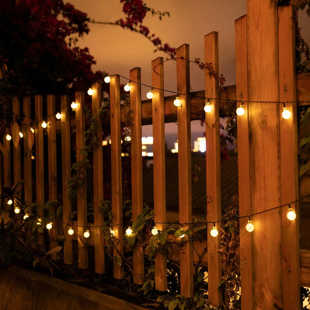 Outdoor wooden fence decorated with warm white solar-powered globe string lights glowing at dusk, creating a cozy garden ambiance with climbing vines and softly blurred evening lights in the background.