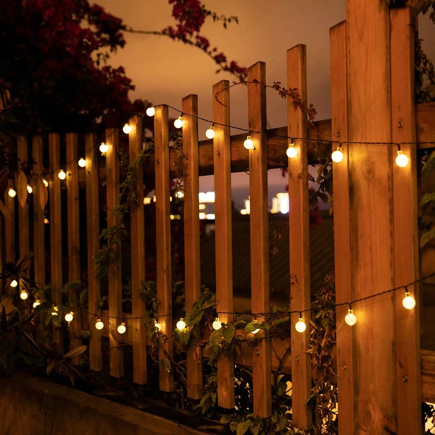 Outdoor wooden fence decorated with warm white solar-powered globe string lights glowing at dusk, creating a cozy garden ambiance with climbing vines and softly blurred evening lights in the background.
